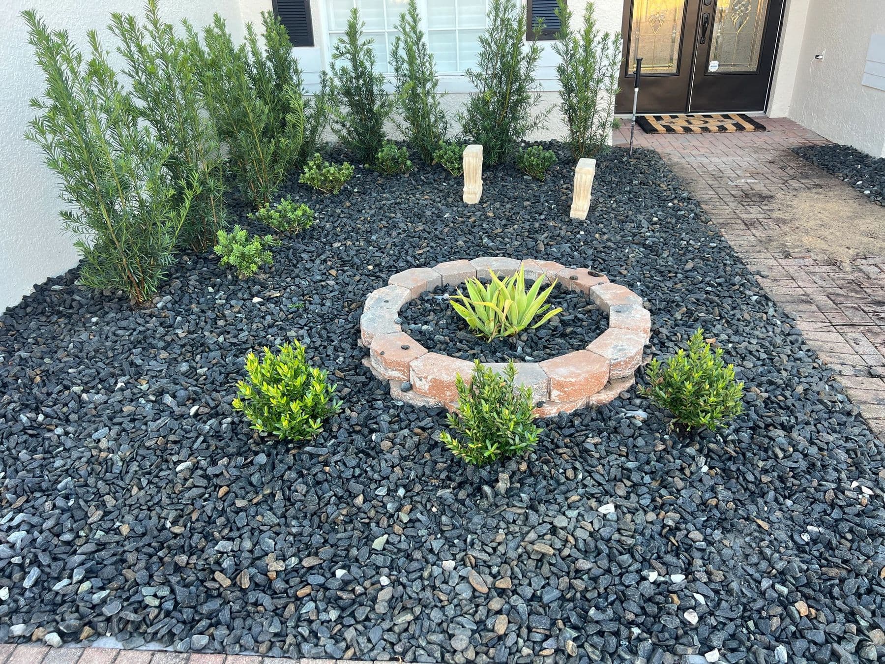 The same entrance bed after work: upright hedges against the wall, dark landscape rock and a circular paver planter ring