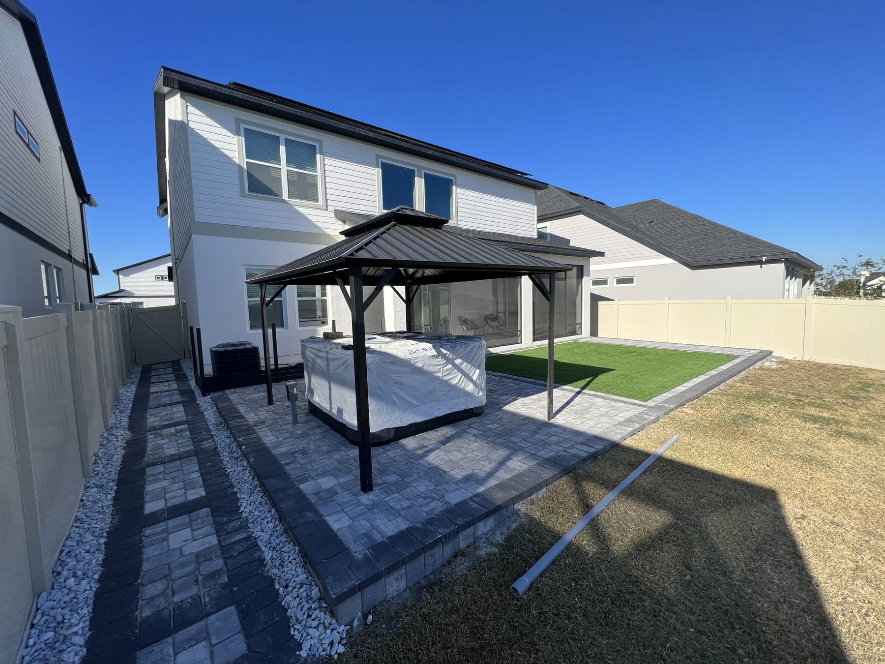 Finished grey paver patio with a dark hardtop pergola over a hot tub, artificial turf to one side and a white stone border along the fence line