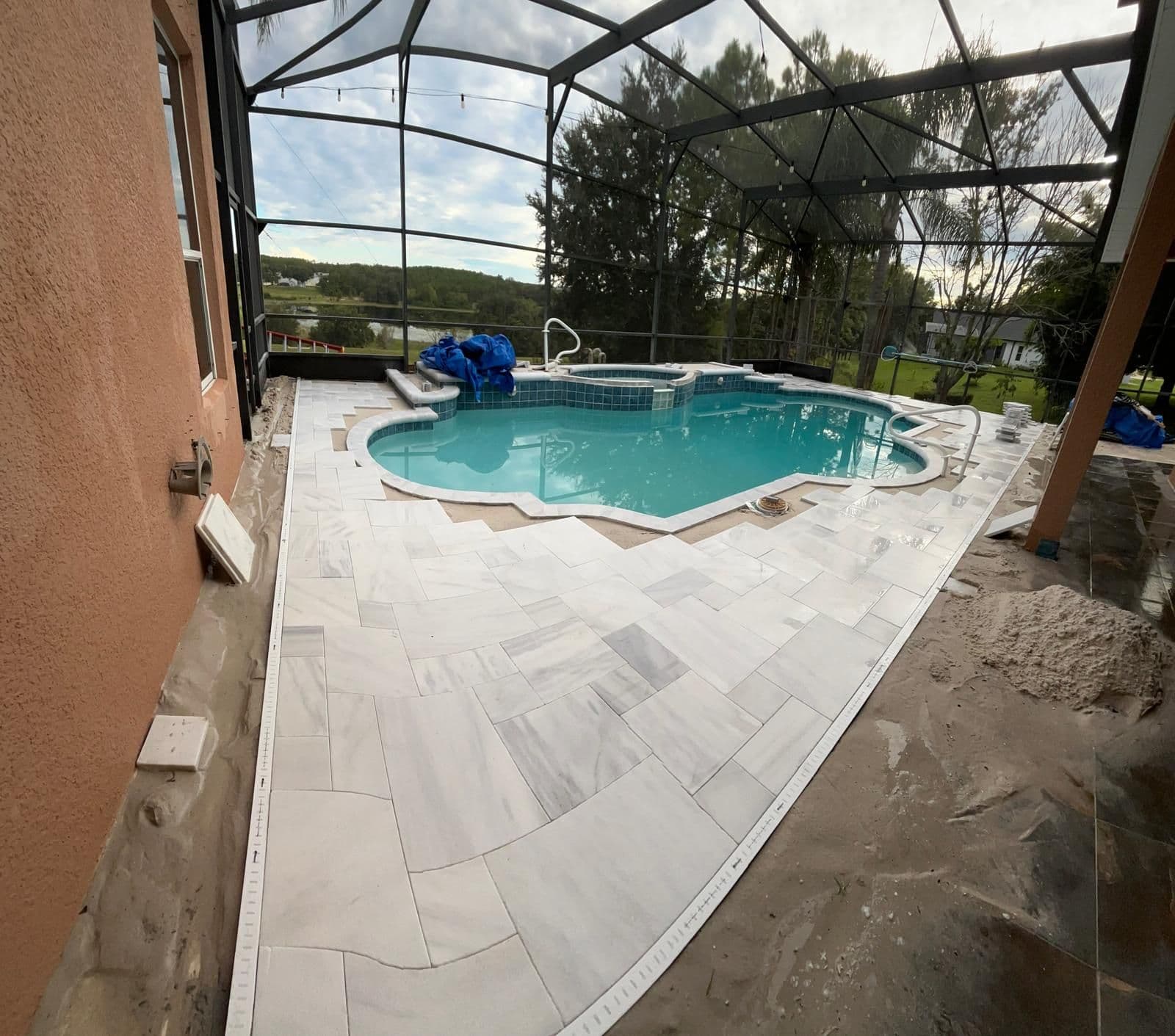 Travertine being laid around the pool inside the screen enclosure, bedding sand visible at the edge