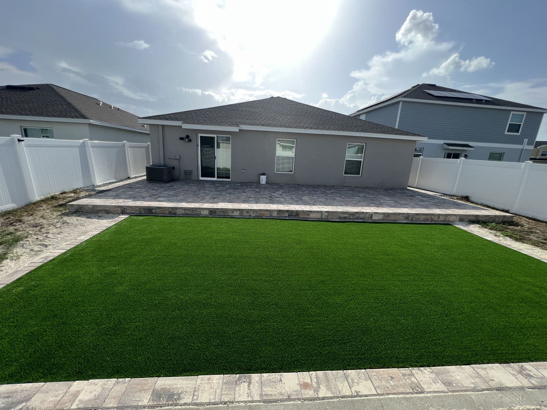 Finished back yard: paver patio running along the house with a large artificial turf lawn in front, enclosed by white fencing