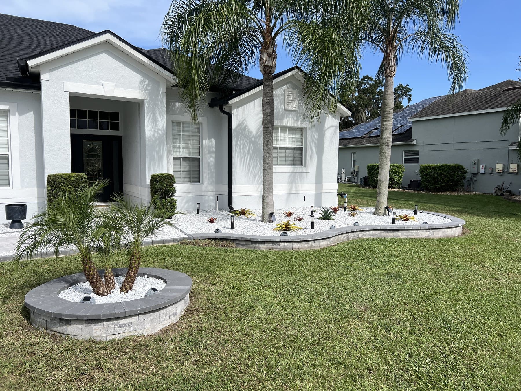 Front yard after work: a curved paver garden wall along the front bed filled with white marble rock and bromeliads, and a circular paver ring around a pygmy date palm on the lawn