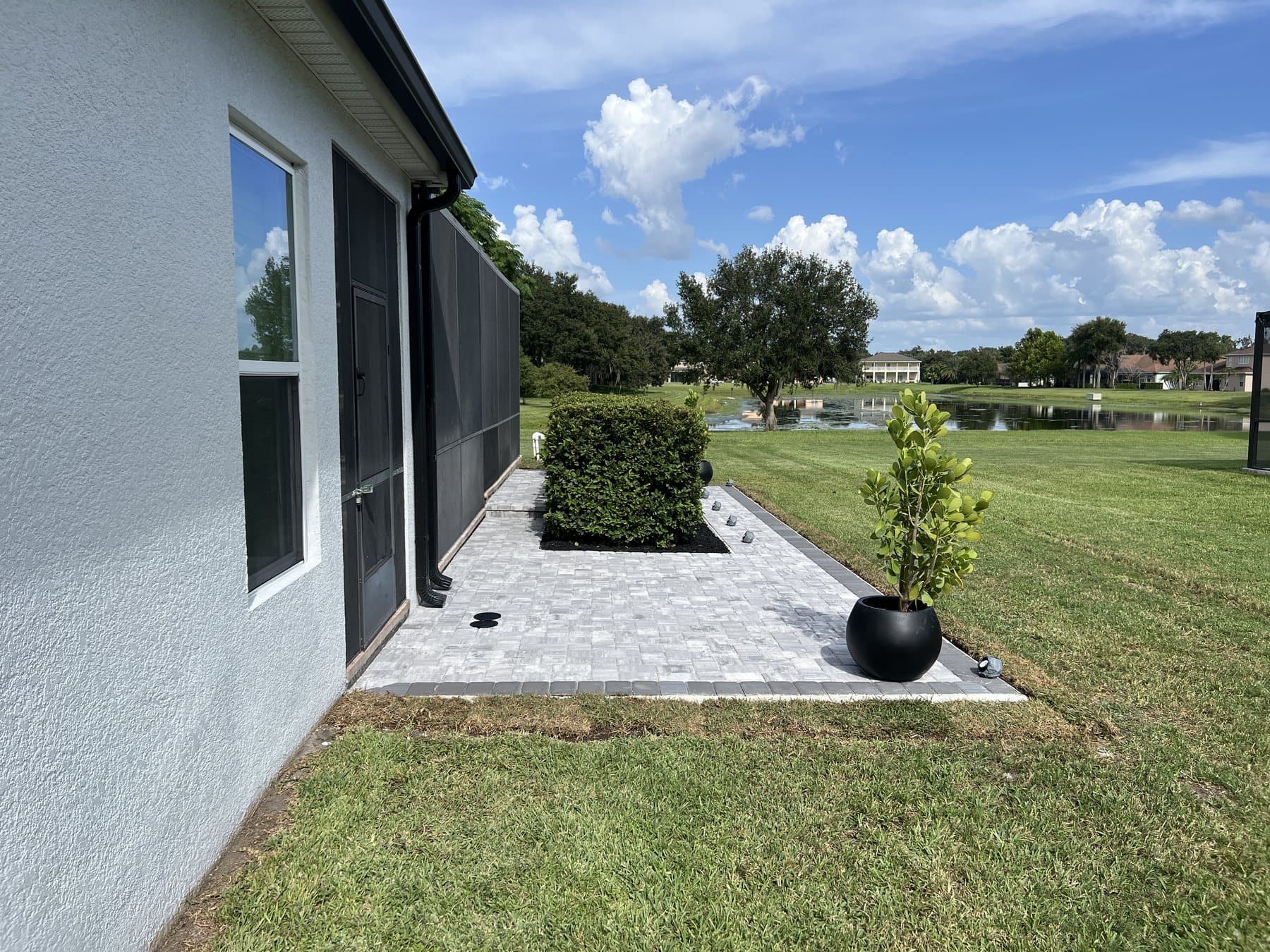 Paver patio running along the house wall past the screen enclosure door, a clipped hedge and a potted clusia in the foreground with a lake beyond