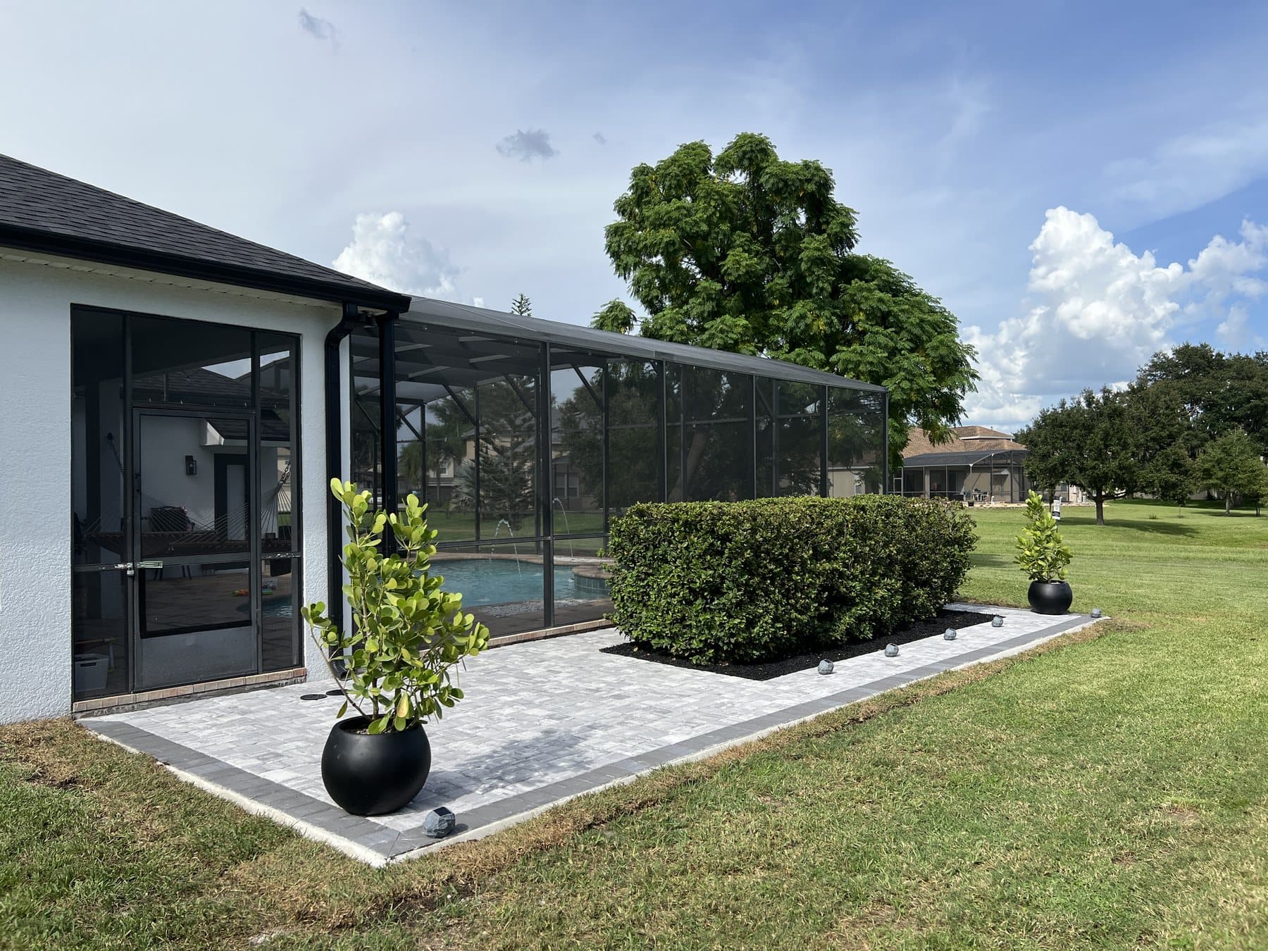 Finished grey paver patio with a charcoal border along the side of a screened pool enclosure, a clipped hedge in a black rock bed and a potted clusia at each end