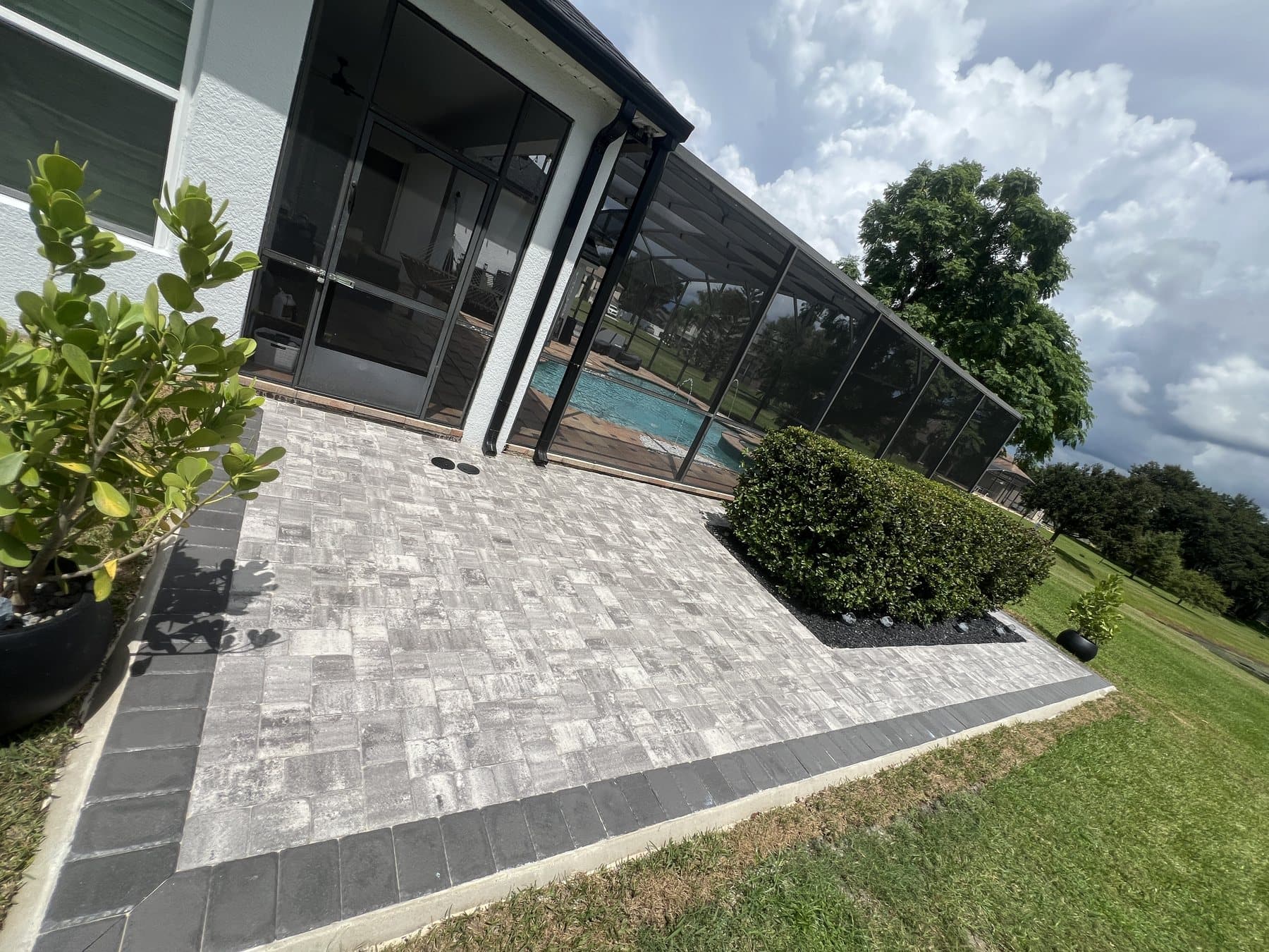 Low angle across the grey paver field to the enclosure, the pool visible through the screen and the hedge to the right