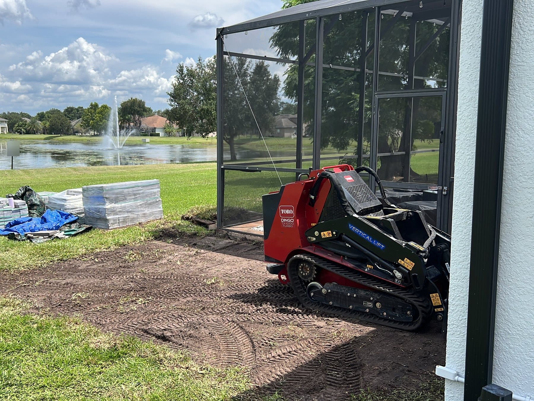 A compact tracked loader working at the corner of the enclosure, pallets of grey pavers on the lawn and the lake fountain behind