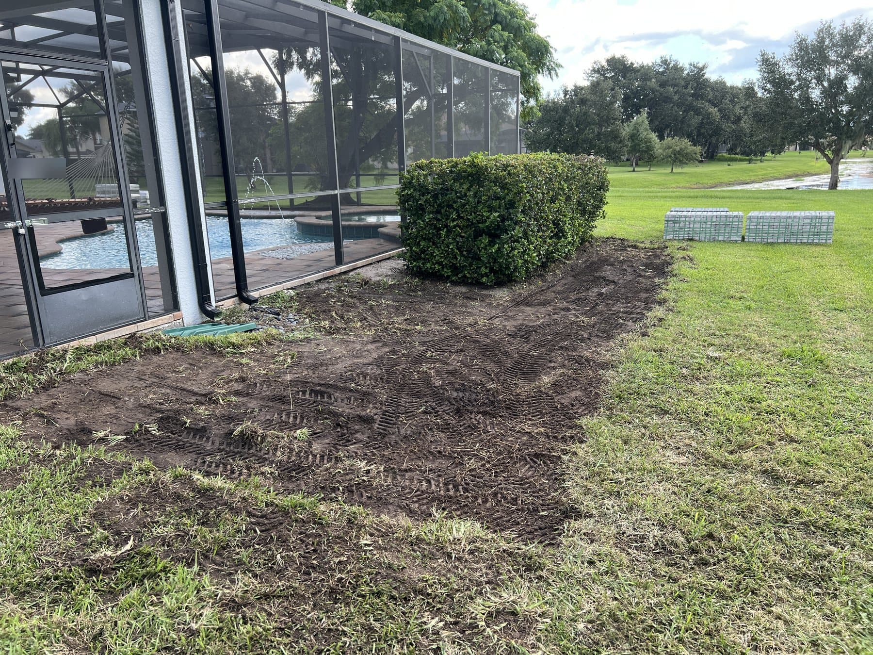 Sod stripped along the enclosure past the hedge, tracked soil exposed, two bundles of pavers stacked on the lawn beyond